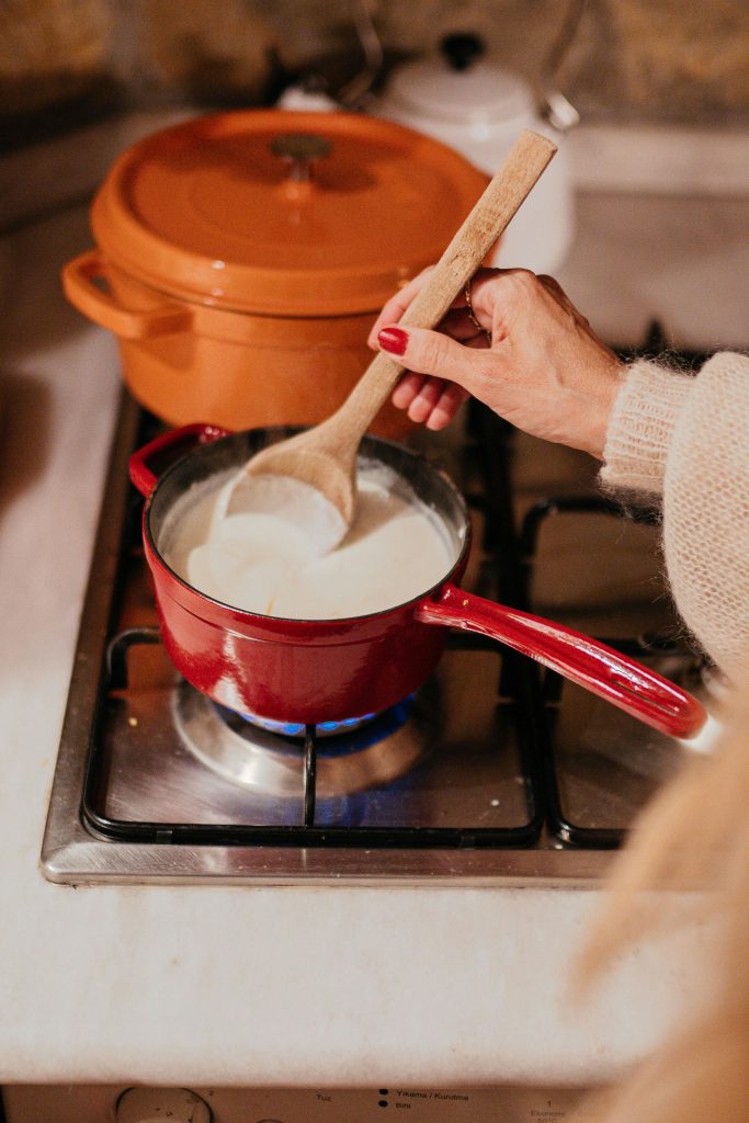 A person stirs milk in a red saucepan on a kitchen stove. Warm, cozy atmosphere.